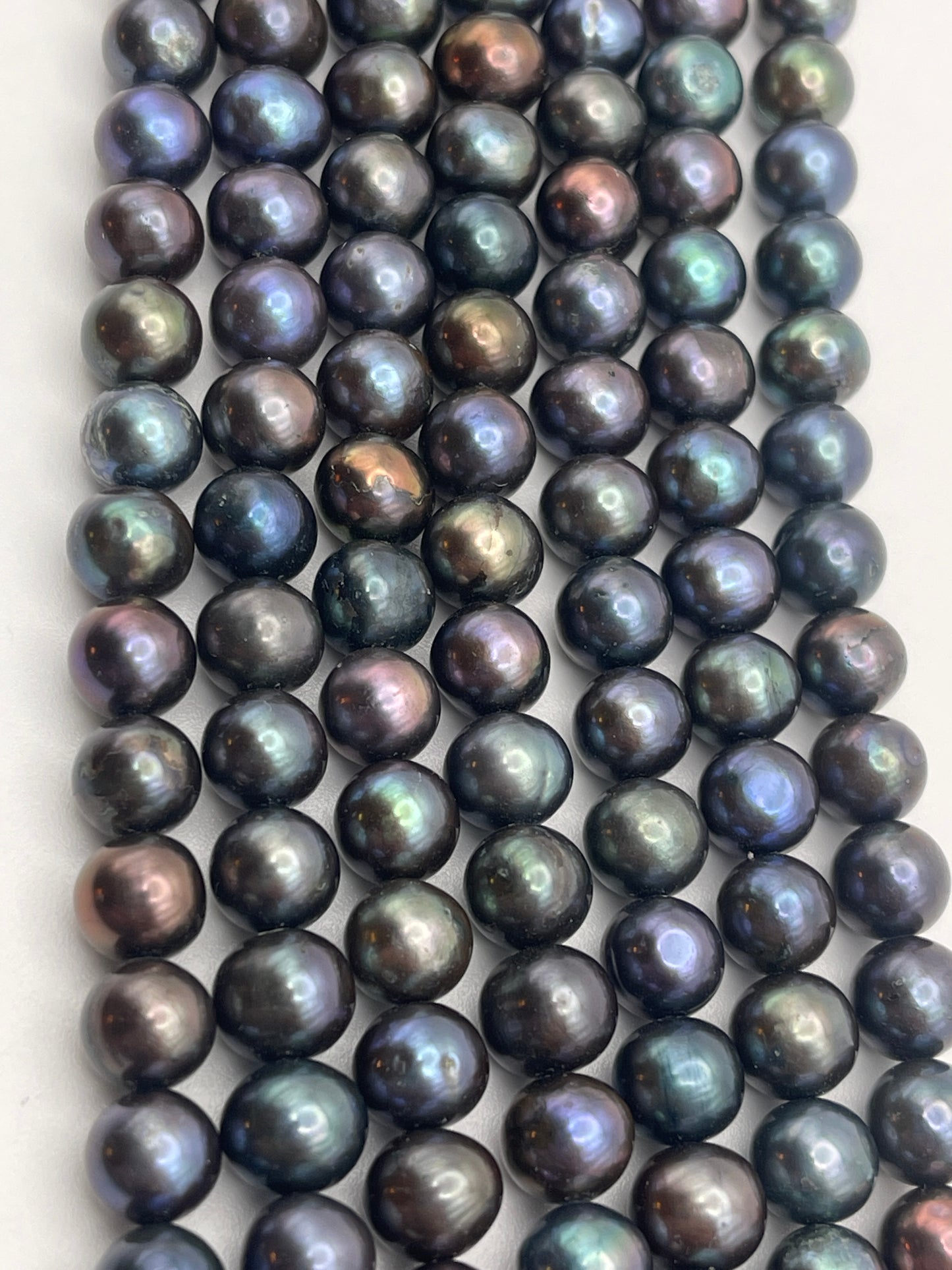 A close-up view of multiple rows of 7-7.5 Peacock potato pearls. The iridescent black pearls display a range of colors including shades of green, blue, purple, and bronze, reflecting light on their smooth, polished surfaces.