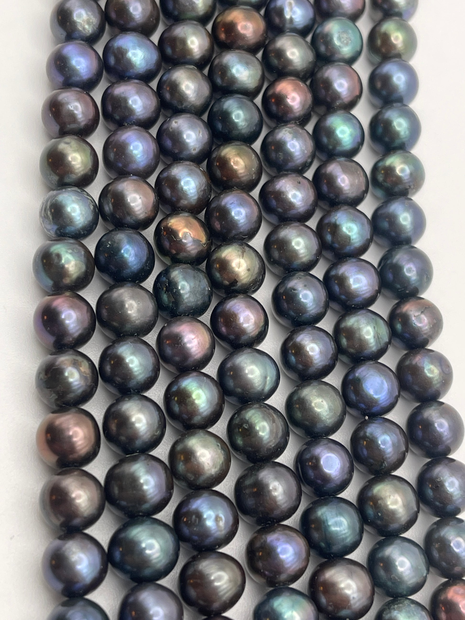 A close-up view of multiple rows of 7-7.5 Peacock potato pearls. The iridescent black pearls display a range of colors including shades of green, blue, purple, and bronze, reflecting light on their smooth, polished surfaces.