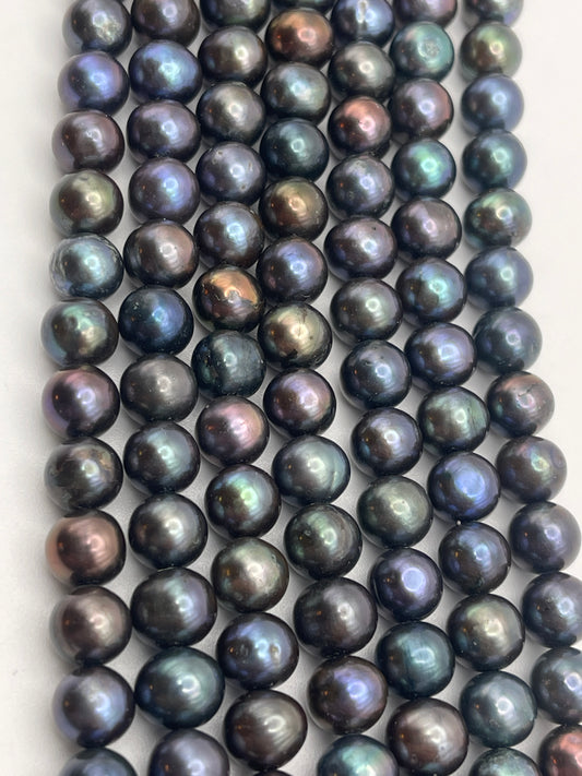 A close-up view of multiple rows of 7-7.5 Peacock potato pearls. The iridescent black pearls display a range of colors including shades of green, blue, purple, and bronze, reflecting light on their smooth, polished surfaces.