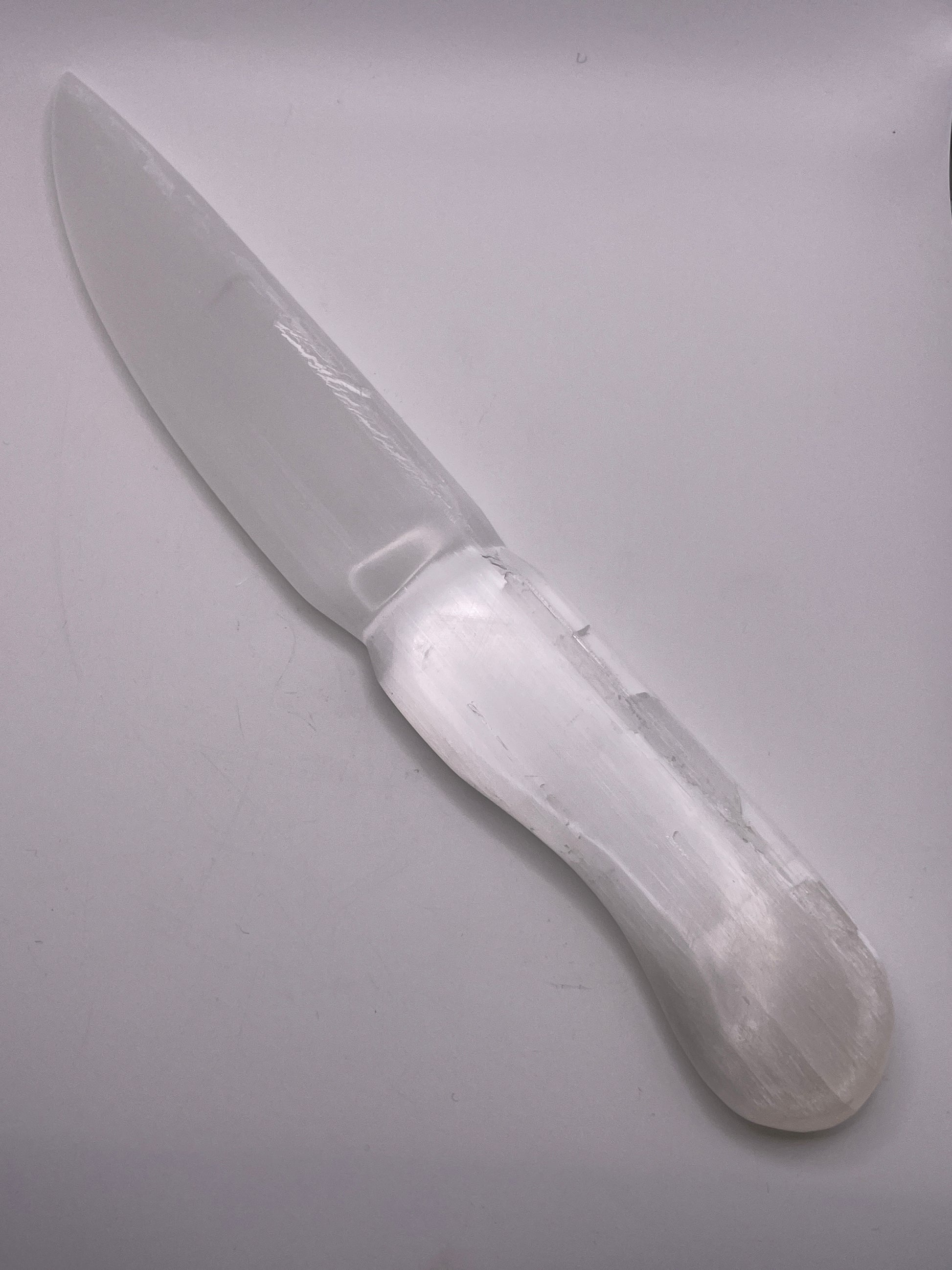 The Selenite Knife 11” is showcased against a plain white backdrop, highlighting its translucent white crystal composition. The knife boasts a smooth, curved blade and a rounded handle, emphasizing the natural texture and sheen of the selenite material.