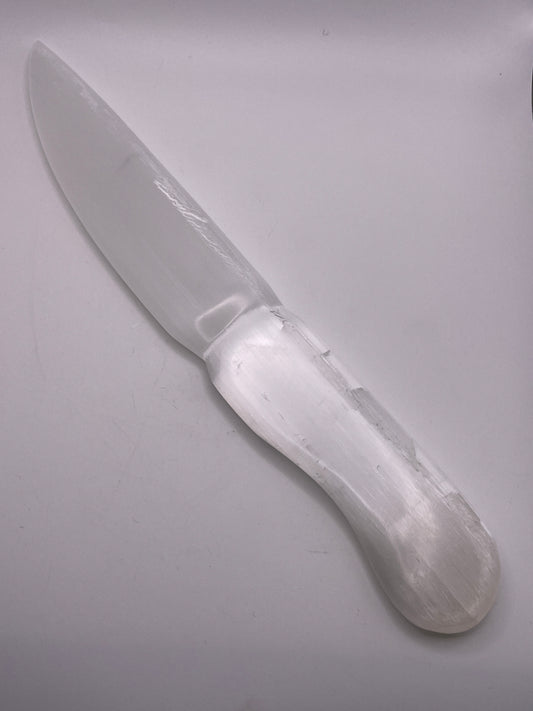 The Selenite Knife 11” is showcased against a plain white backdrop, highlighting its translucent white crystal composition. The knife boasts a smooth, curved blade and a rounded handle, emphasizing the natural texture and sheen of the selenite material.