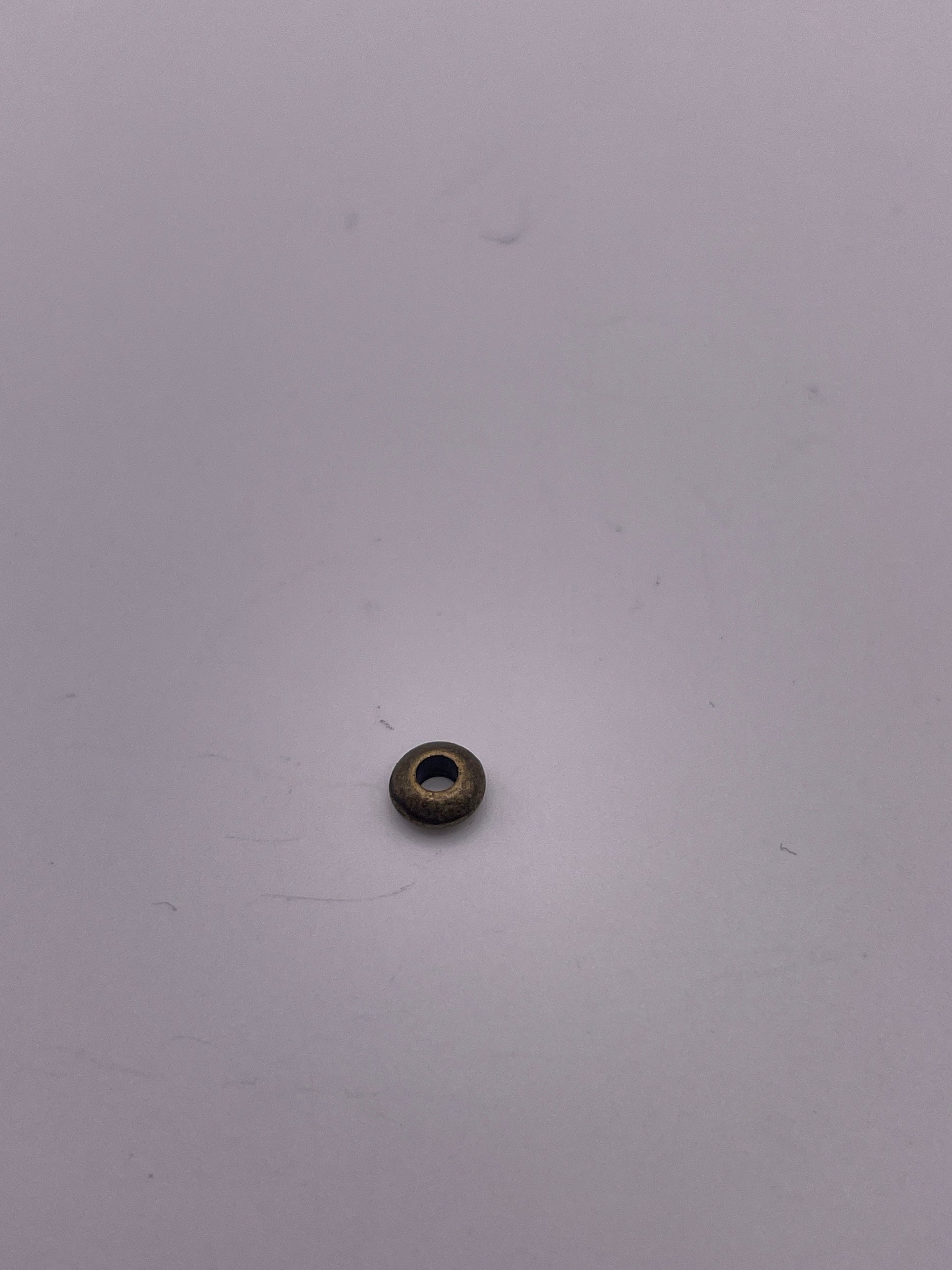 A Pewter Rondelle .35, a small, round bead with a hole in the center, sits on a white surface. The bead has a metallic finish and the lighting casts a faint shadow around it.