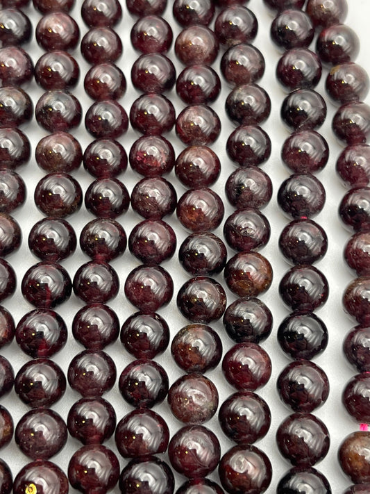 Close-up image of several rows of 8mm Garnet Rnd Smooth beads arranged on a flat surface. The polished, dark red beads have a glossy finish, reflecting light and showing variations in color intensity.