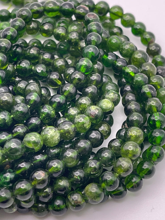 A close-up view of multiple strands of 5mm Chrome Diopside Rnd beads with varying shades of green. The beads have a glossy finish, and some appear slightly translucent, displaying subtle inner patterns. They are arranged in a loose, spiraled pile on a white background.