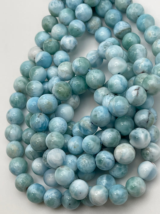 A close-up view of the "8mm Larimar Rnd Smooth" necklace, showcasing round, polished beads with varying shades of blue—from light to deep blue—and adorned with white and greenish marbling. The necklace is elegantly coiled in a loose loop on a white surface.