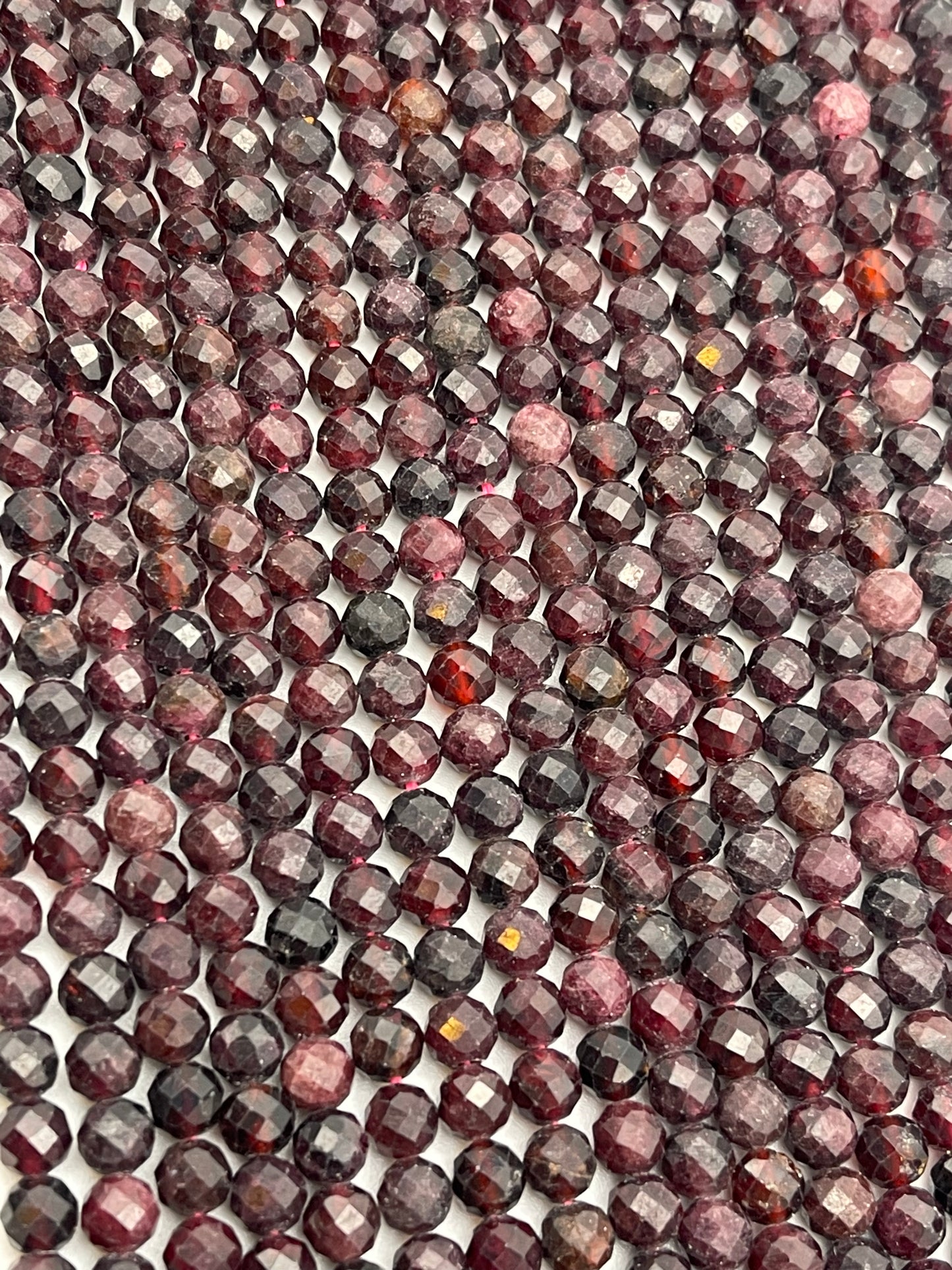 The 5mm Fac Garnet Rnd beads, in various shades of burgundy, red, and dark purple, are tightly lined up next to each other, filling the entire frame. Their polished surfaces create a shimmering, repetitive pattern.