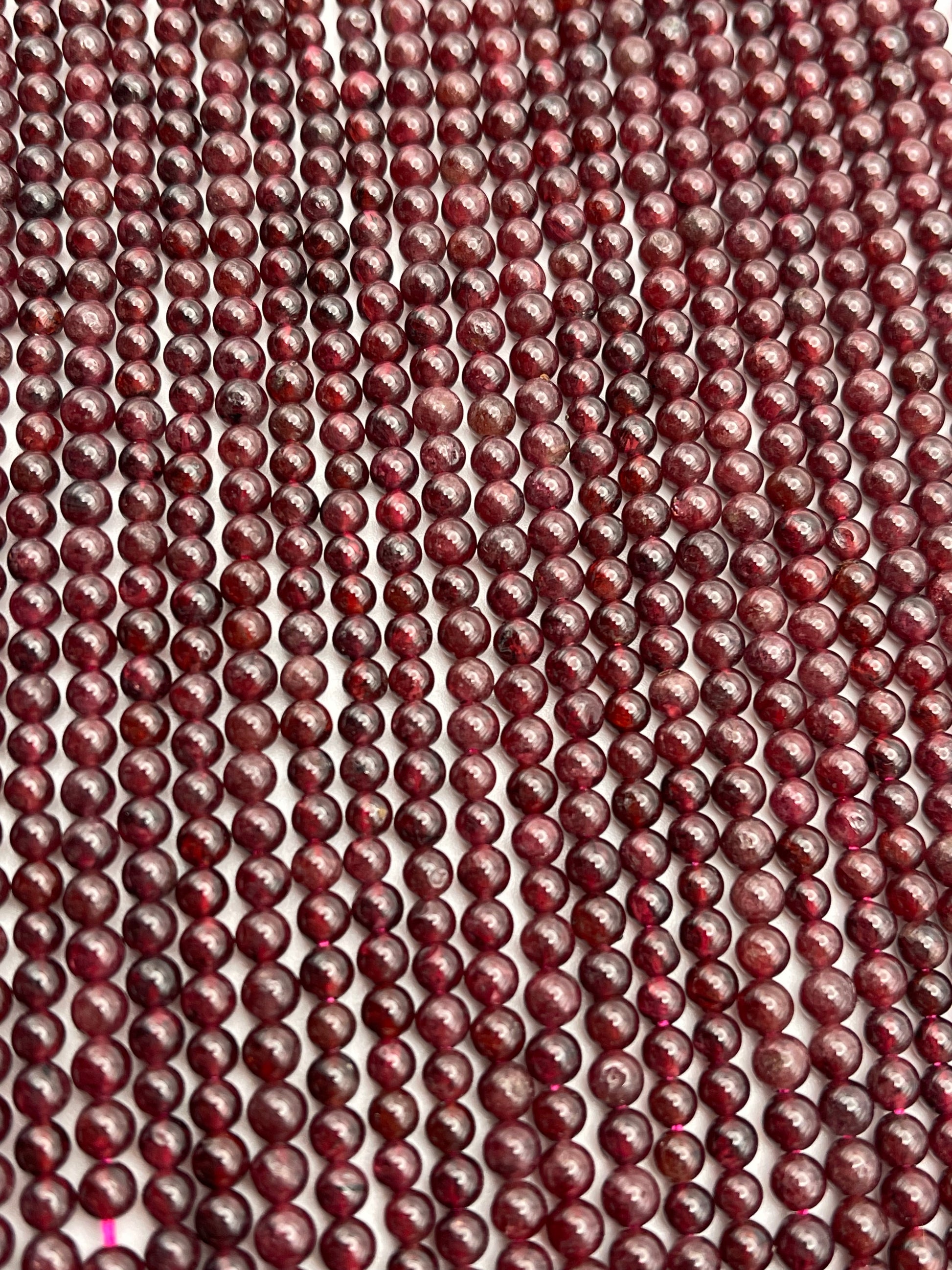 Close-up view of numerous 4mm Round Garnet beads aligned in parallel rows, creating a textured and pattern-rich visual. The surface of each garnet bead exhibits slight variations in color and sheen, adding to the intricate detail of the arrangement.