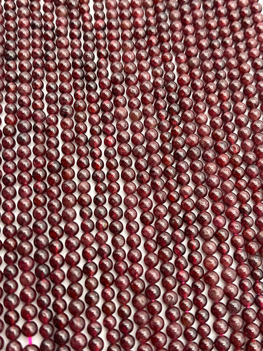 Close-up view of numerous 4mm Round Garnet beads aligned in parallel rows, creating a textured and pattern-rich visual. The surface of each garnet bead exhibits slight variations in color and sheen, adding to the intricate detail of the arrangement.