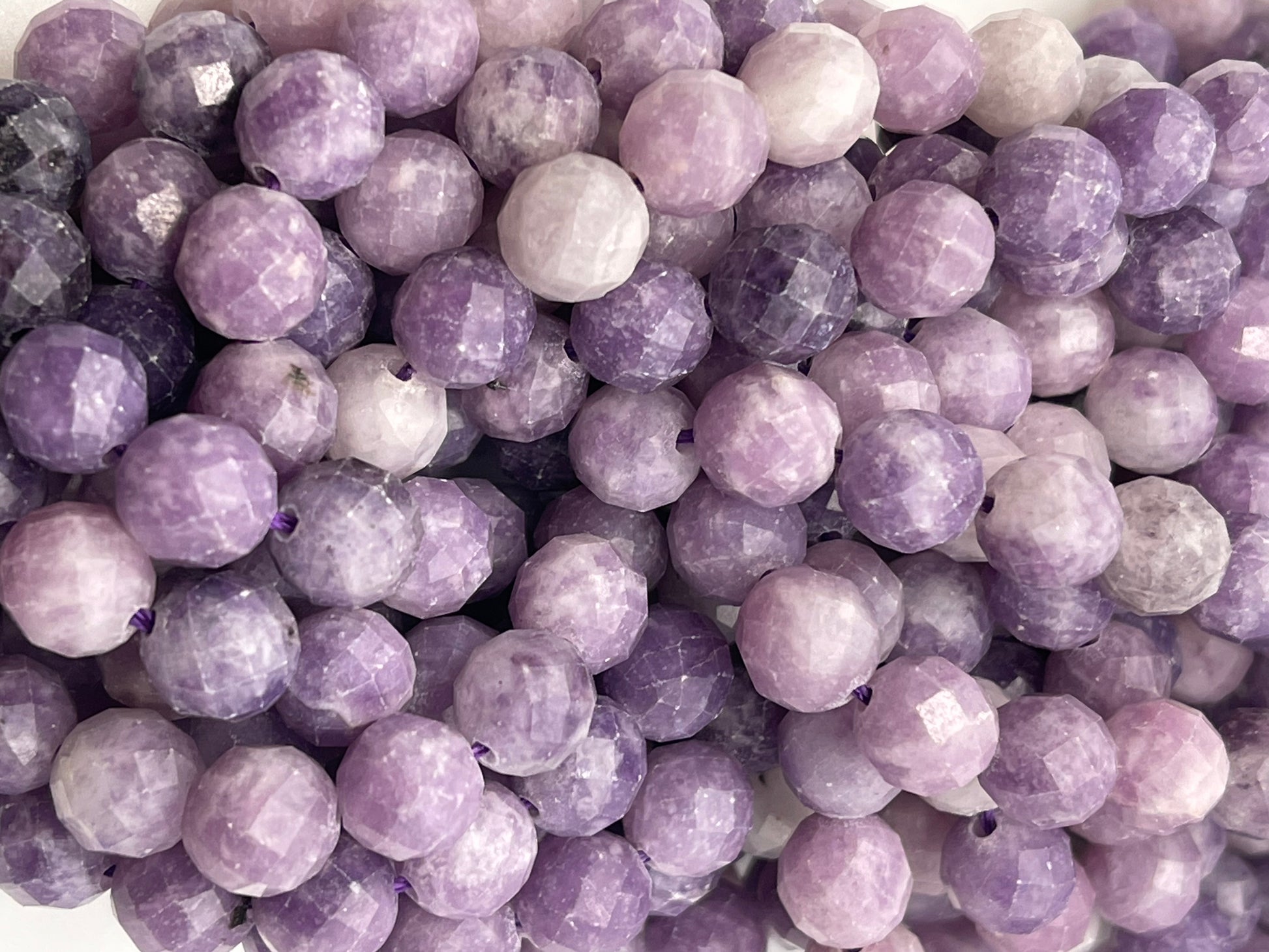 A close-up view of multiple 8mm Faceted Lepidolite Round gemstone beads. The beads showcase various shades of purple, ranging from light lavender to deep purple, and are strung closely together, highlighting their shiny and polished surfaces.