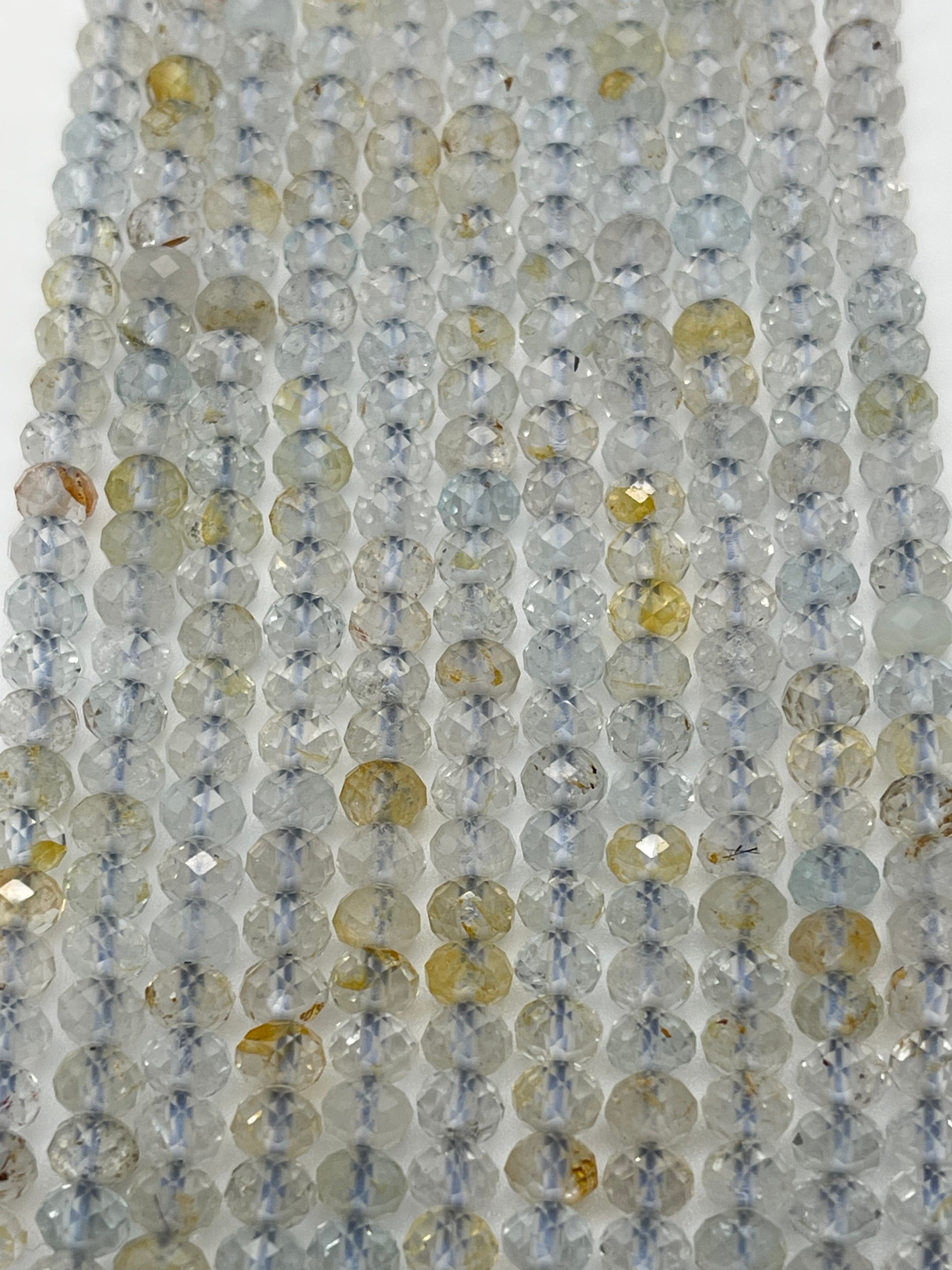 Strings of 5mm Fac Topaz Rondell beads in an array of light gray and yellow hues are strung closely together. The beads have a translucent quality with some displaying flecks of brown and other colors, creating a shimmering effect under the light.