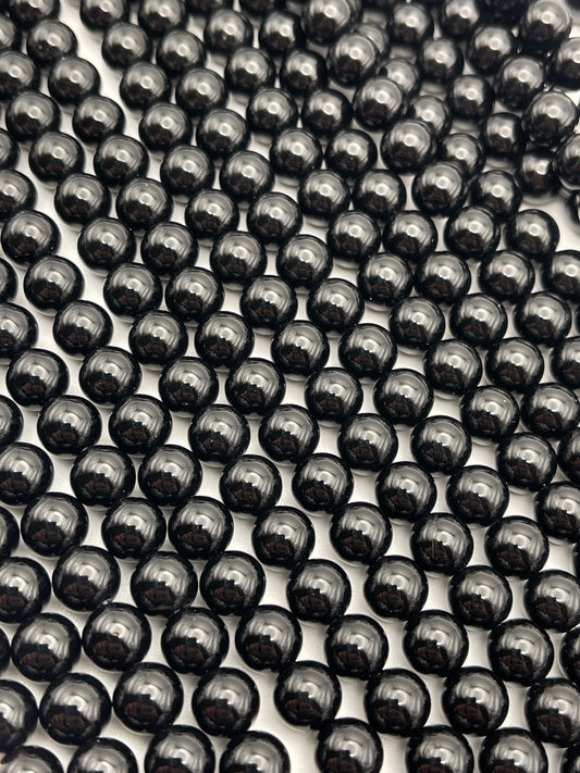 A close-up image of numerous 8mm Black Onyx beads arranged in a grid pattern. The beads reflect light, giving them a polished and glossy appearance. The background is out of focus, highlighting the uniformity and alignment of the 8mm Black Onyx beads.