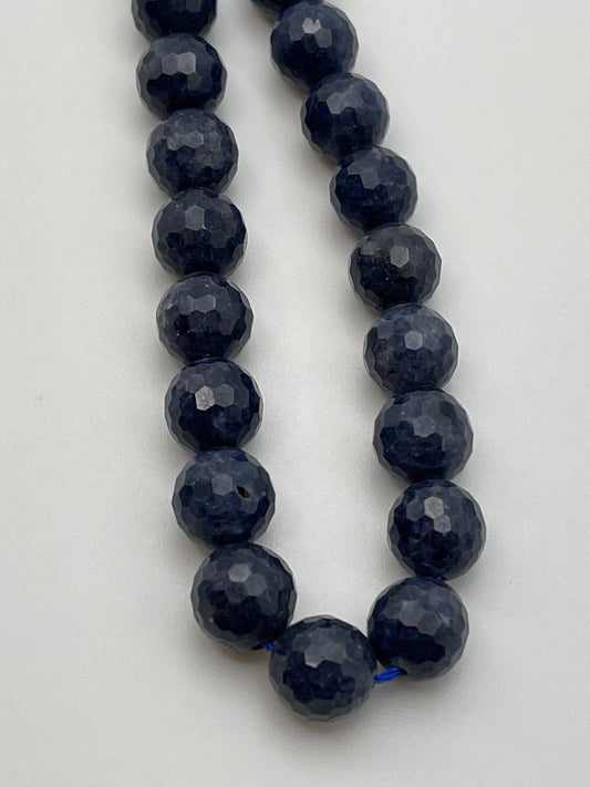 A close-up image showcasing a strand of 8mm Fac Sapphire Rnd Blue beads, arranged linearly against a plain white background. The dark blue, faceted round beads appear uniformly sized and exhibit a glossy finish.