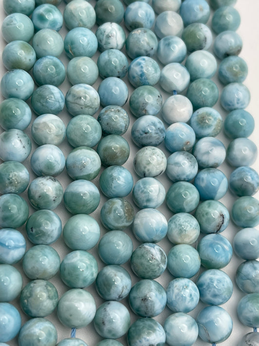 The image shows multiple strands of 8mm Round Larimar beads in various shades of blue and green. The beads are closely packed together and display a marbled pattern with subtle variations in color and texture, ranging from light to dark hues.