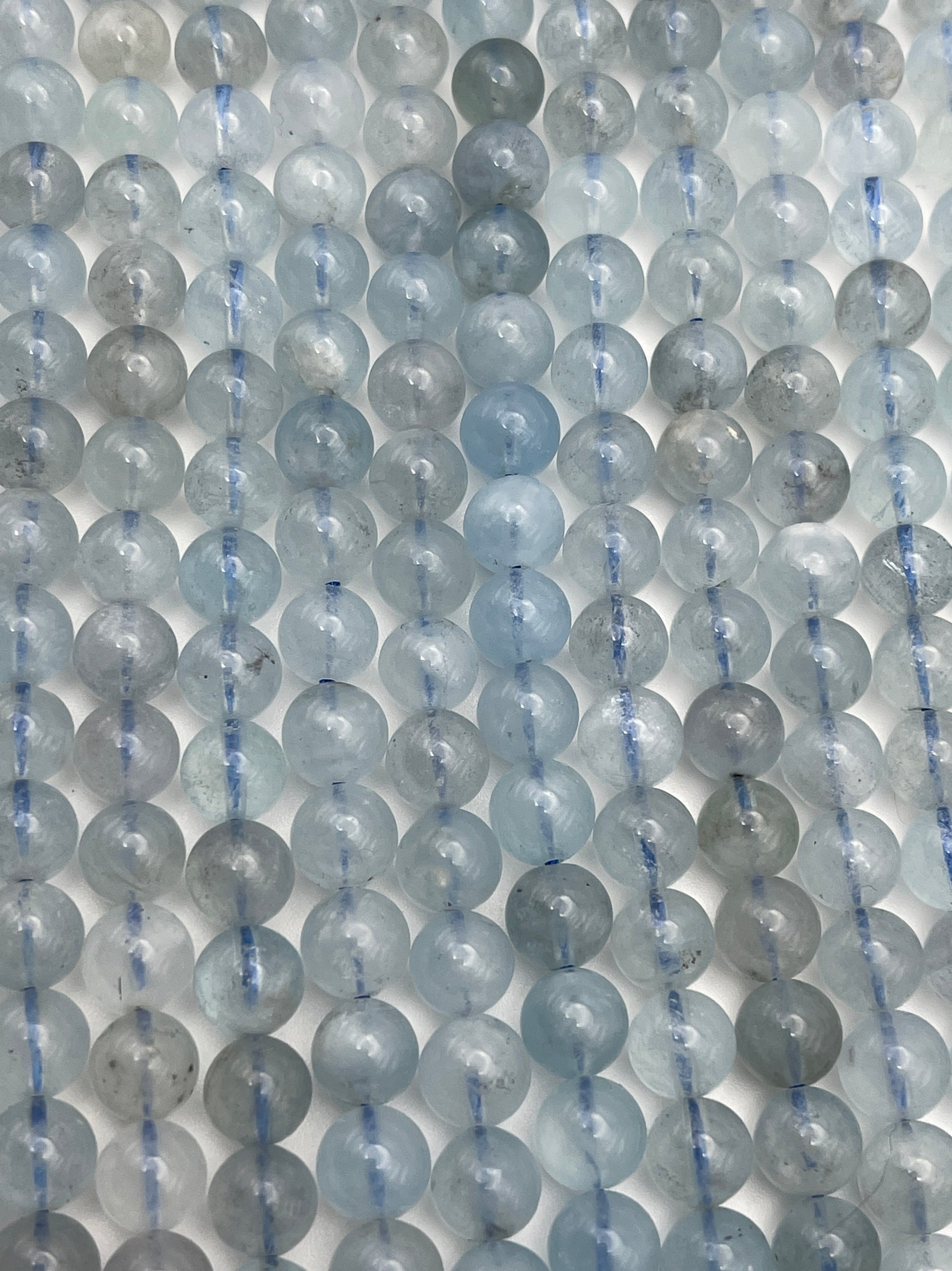 Rows of 8mm Round Aquamarine beads, in translucent blue and gray, are strung closely together to form a repetitive pattern. Each bead has a slightly shiny surface with subtle color variations. The strings connecting the beads are barely visible.