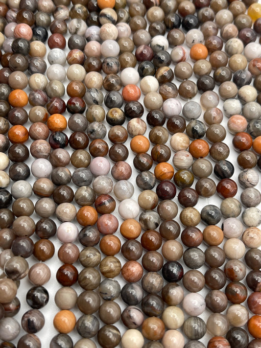 A collection of 6mm Round Petrified Wood beads in various earthy tones, including shades of brown, beige, gray, white, and red. The beads are arranged closely together, creating a textured pattern on a flat surface.