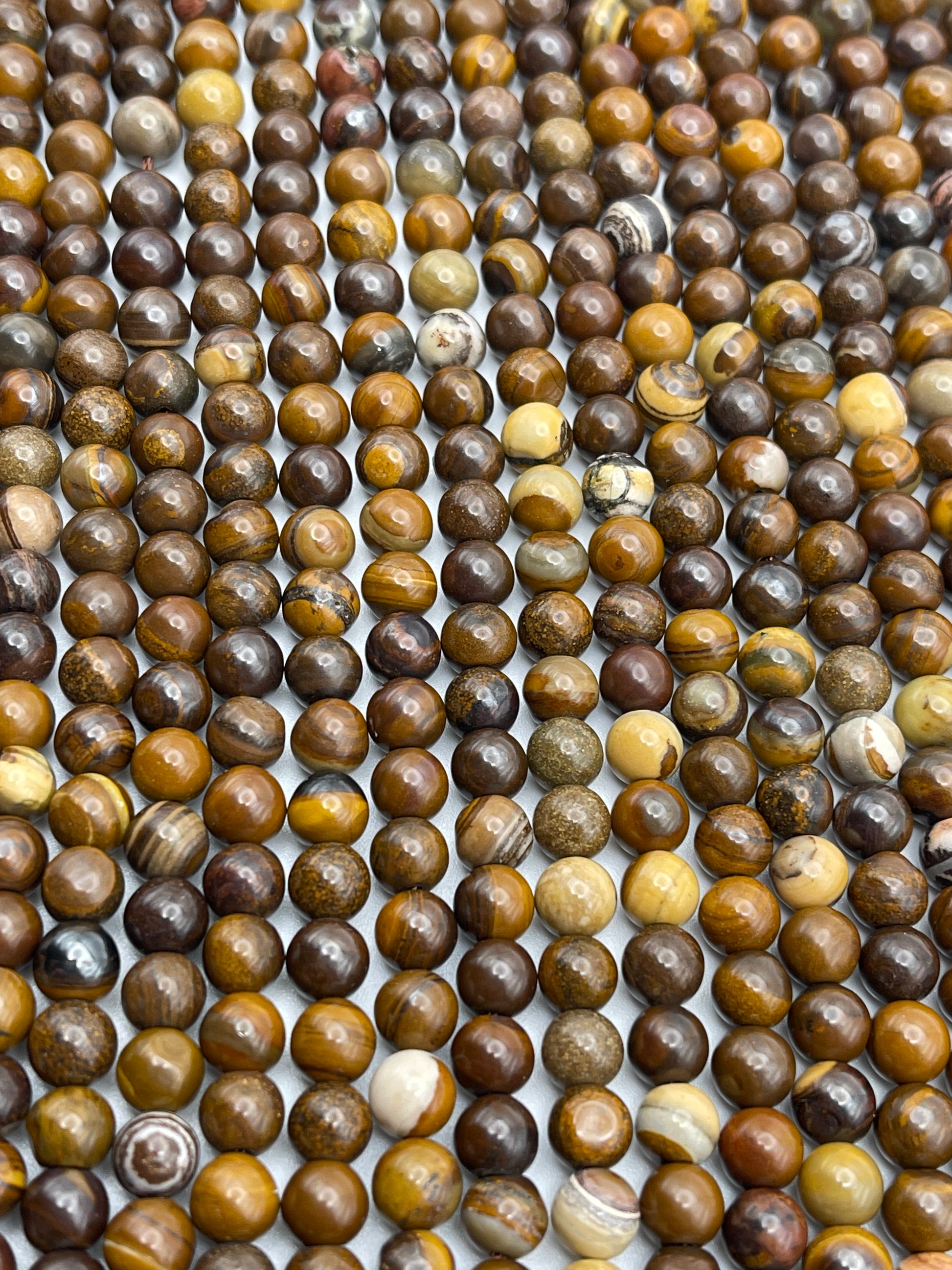 Rows of 6mm Tiger Iron Round Jasper beads, polished in shades of brown, yellow, and beige, are laid out on a flat surface. The slight variations in color and pattern among the beads give the arrangement a natural, earthy appearance.