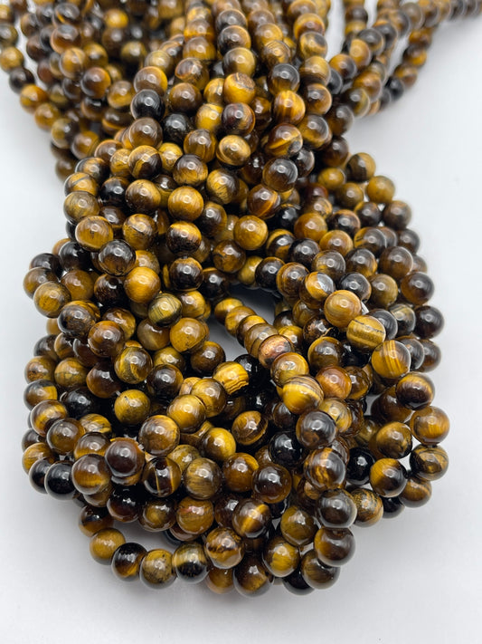Close-up image of multiple strands of 6mm Tiger's Eye Round beads. The spherical beads exhibit a blend of rich golden-brown, deep brown, and dark yellow hues, creating the characteristic shimmering effect typical of tiger's eye stones. The beads are closely arranged and reflect light beautifully.