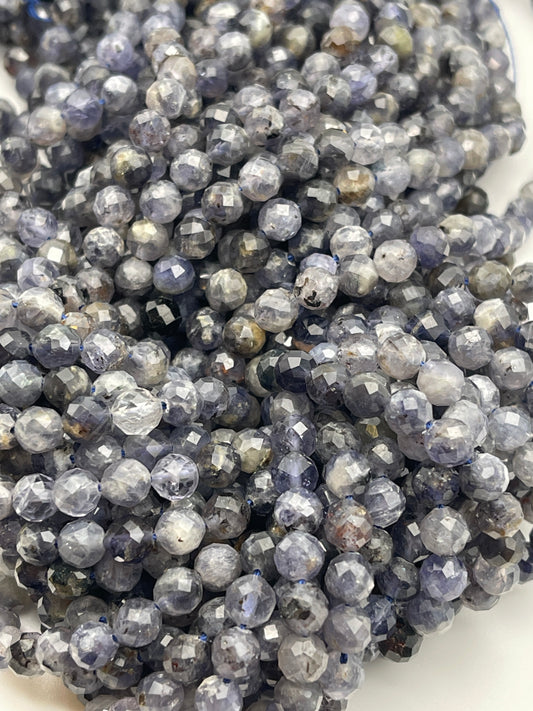 A close-up view of numerous small, round, 6mm Fac Iolite Rnd PG beads in shades of grey, blue, and light brown. The beads are arranged tightly together, creating a textured and shimmering surface.