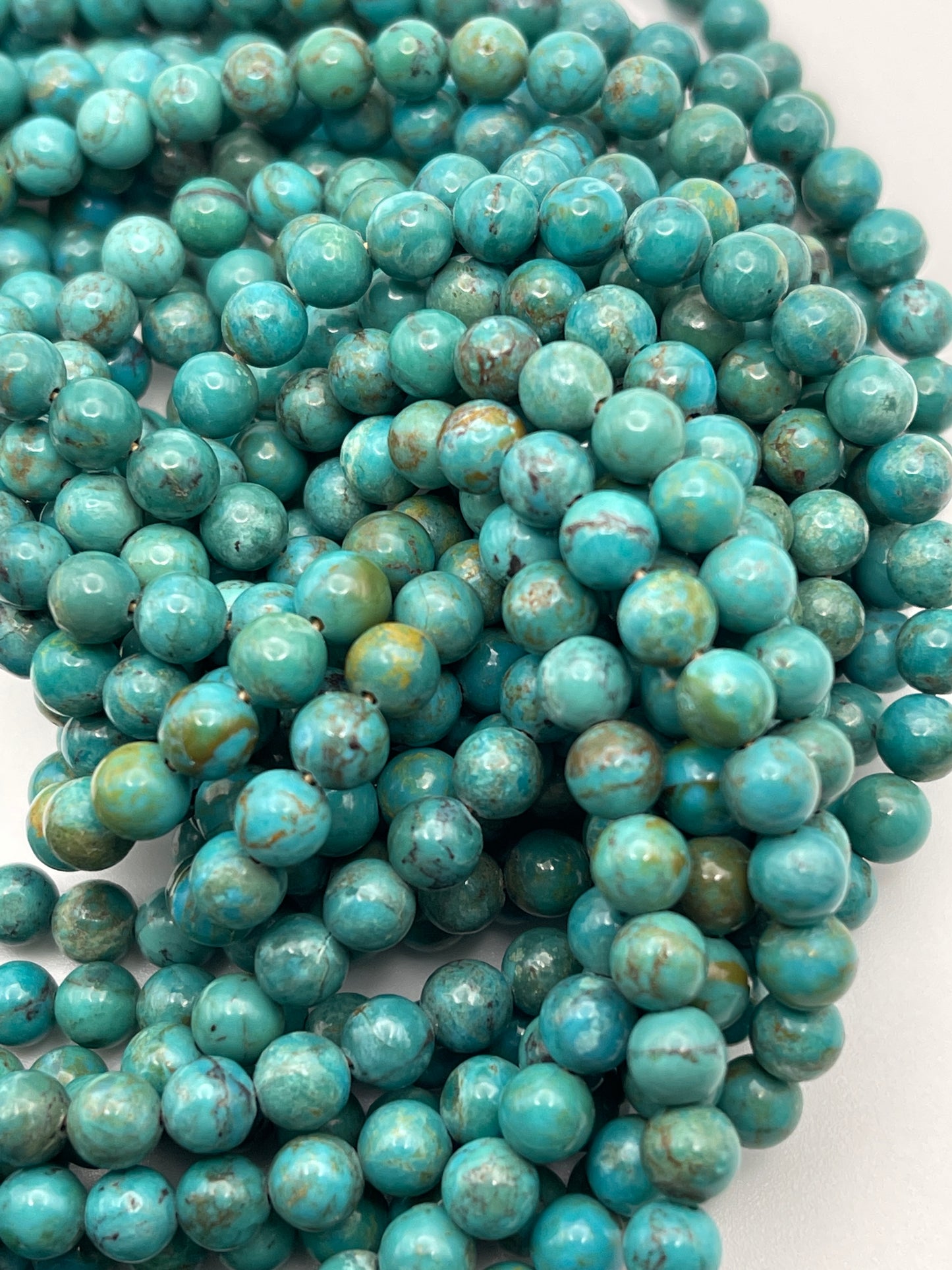 A close-up image of multiple strands of 6mm Turquoise Rnd A beads. The beads vary slightly in color, displaying shades of blue and green, and feature a marbled texture with natural veining. The intertwined strands create a textured, layered effect.