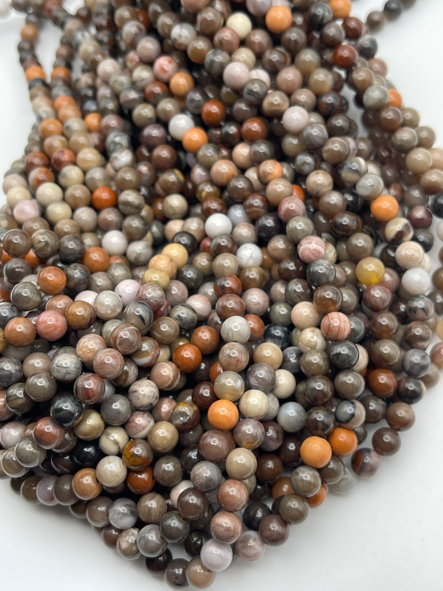 Close-up image of multiple strands of 6mm Round Petrified Wood beads in various earthy tones, including shades of brown, gray, pink, orange, and beige, arranged in a slightly tangled cluster on a white background.
