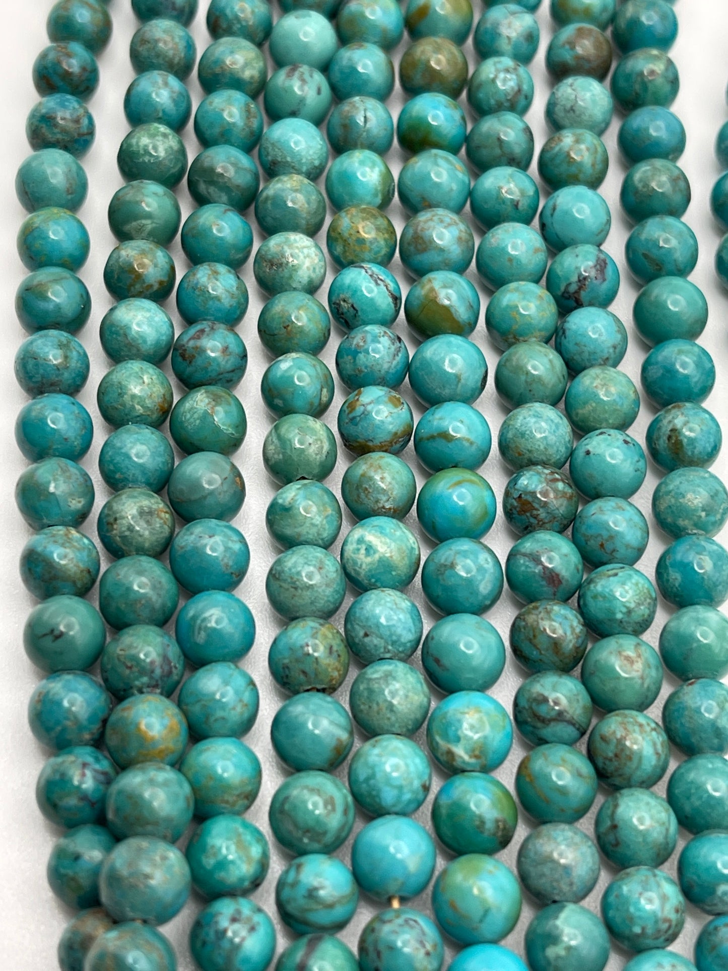 Rows of 6mm Turquoise Rnd A beads are neatly arranged on a flat surface. The smooth, round beads vary slightly in hue, showcasing different shades of blue and green, with some featuring subtle brownish veining. The overall appearance is colorful and visually striking.