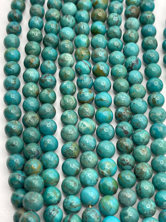 Rows of 6mm Turquoise Rnd A beads are neatly arranged on a flat surface. The smooth, round beads vary slightly in hue, showcasing different shades of blue and green, with some featuring subtle brownish veining. The overall appearance is colorful and visually striking.
