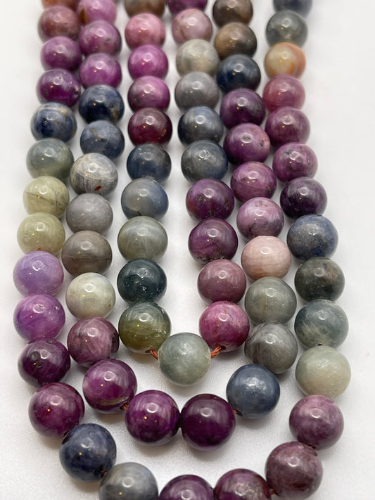 A close-up of several strands of 8mm Sapphire Rainbow Rnd Smooth beads in shades of purple, pink, blue, and green. The beads are glossy with a smooth texture, displaying natural variations in color and pattern. The strands are arranged parallel on a white surface.
