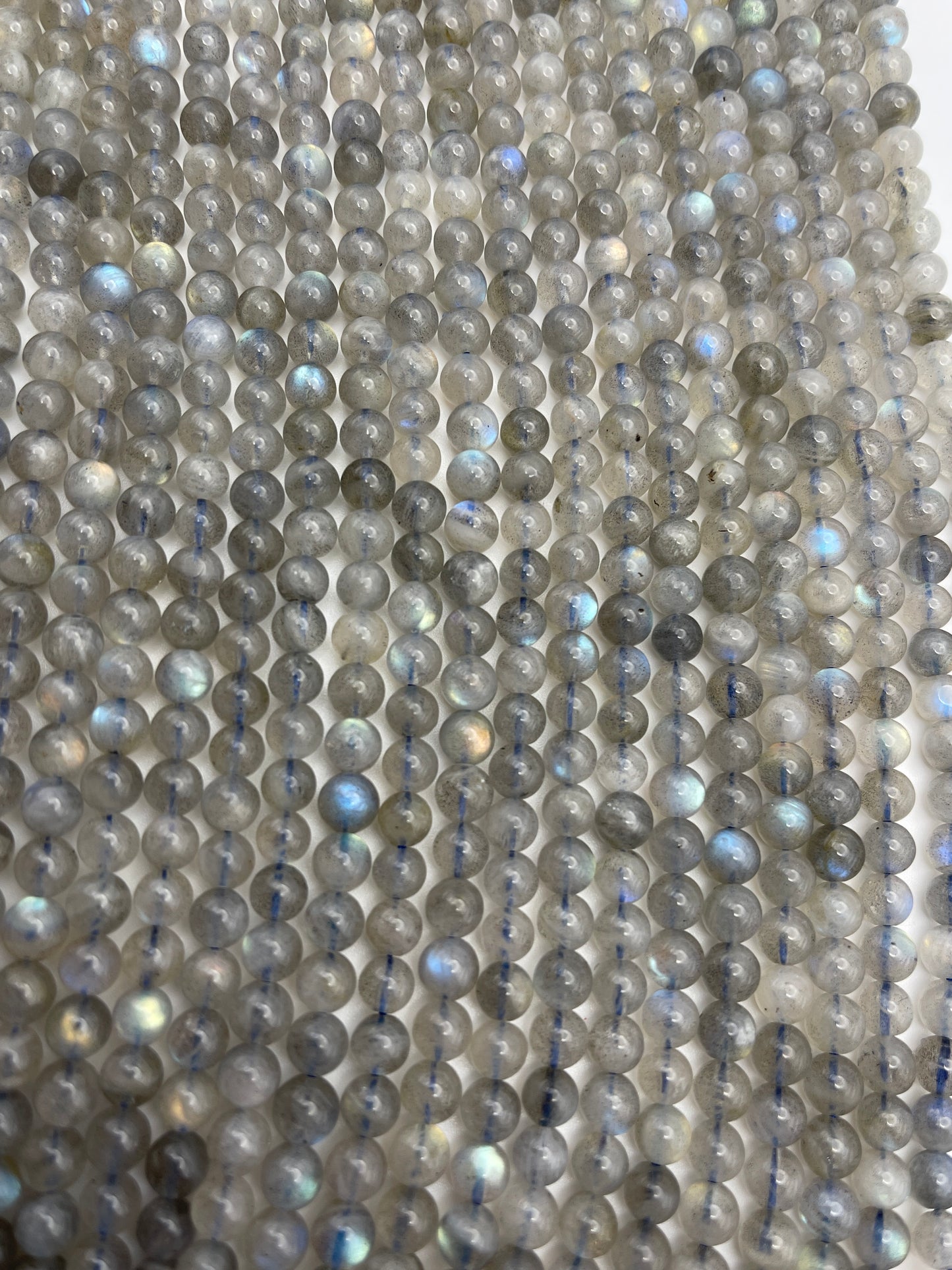 A close-up view of multiple strands featuring the 6mm Round Labradorite beads with a greyish hue and occasional iridescent flashes, laid out parallel to each other.