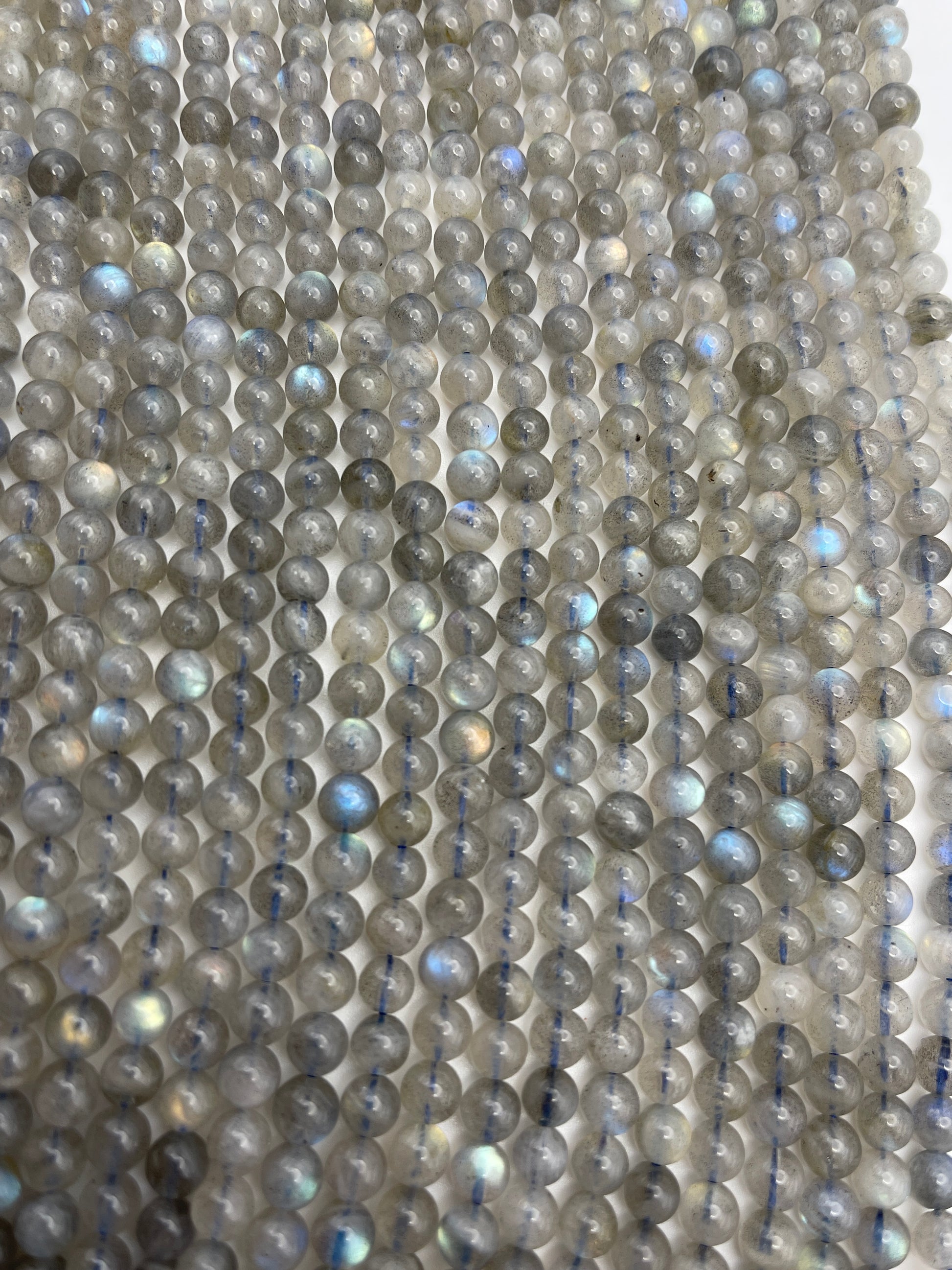 A close-up view of multiple strands featuring the 6mm Round Labradorite beads with a greyish hue and occasional iridescent flashes, laid out parallel to each other.