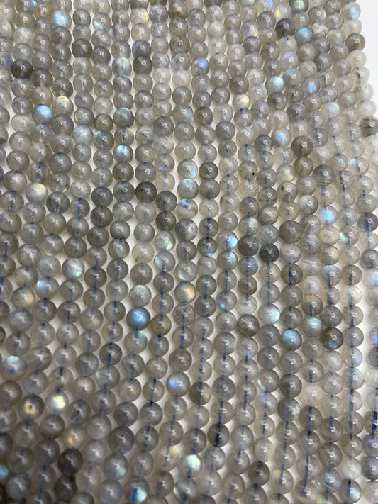 A close-up view of multiple strands featuring the 6mm Round Labradorite beads with a greyish hue and occasional iridescent flashes, laid out parallel to each other.