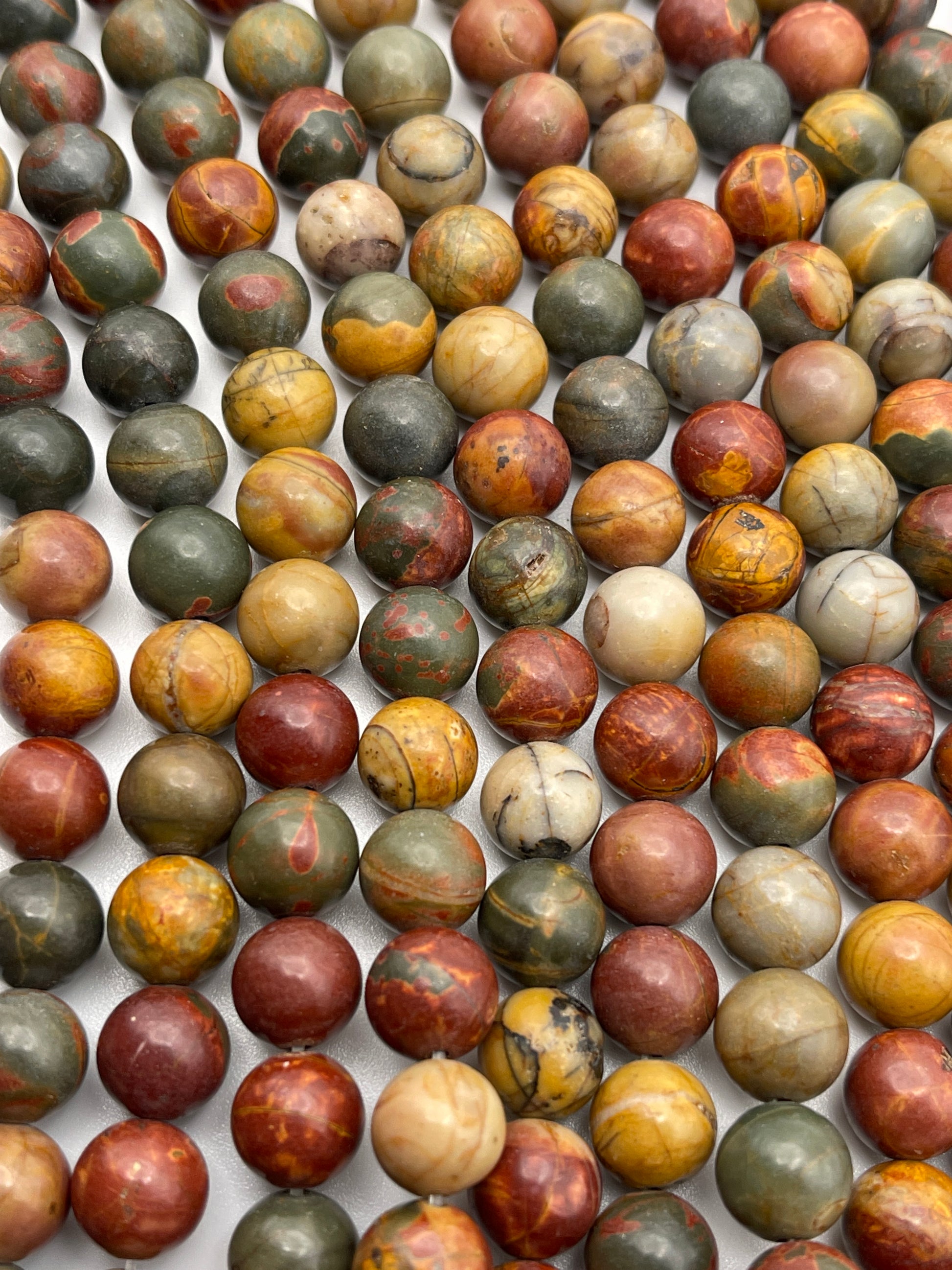 Close-up of a collection of 8mm Red Creek Round Jasper beads in various earthy tones, including shades of green, brown, red, yellow, and cream, arranged in rows. The beads have a smooth surface with visible natural patterns, giving them a marbled appearance.