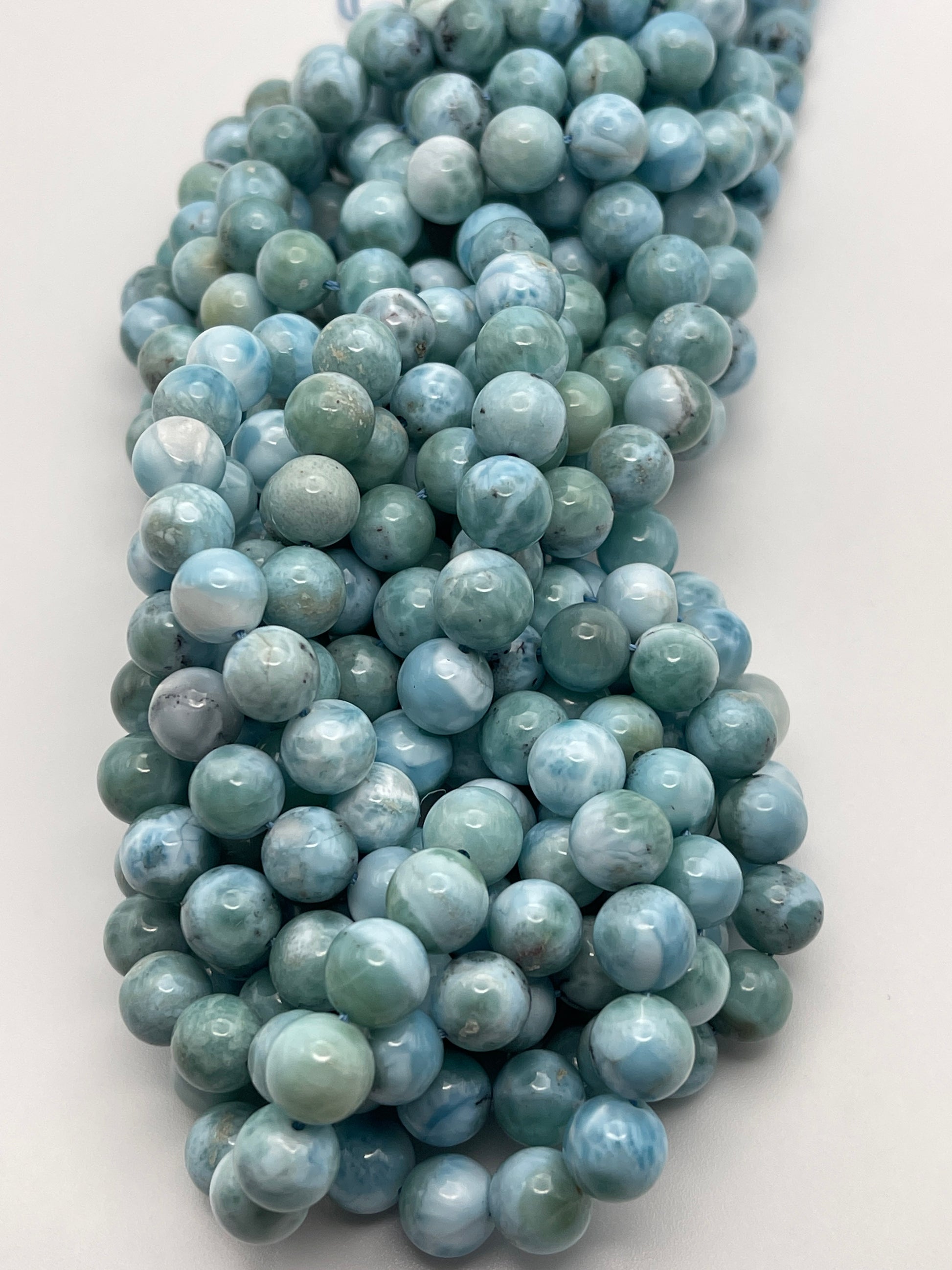 A bundle of 8mm Round Larimar beads in various shades of light blue and green is loosely coiled on a white surface. The beads have a glossy finish, showcasing their natural patterns and marbling.