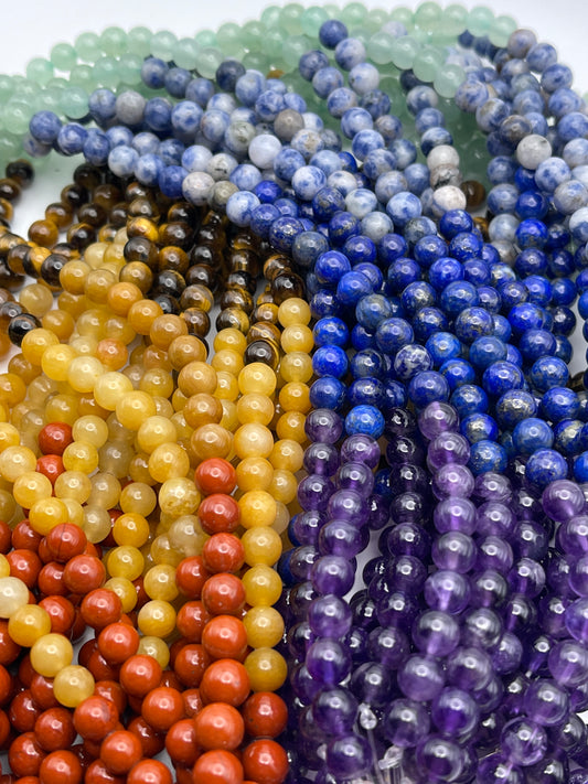 A close-up image featuring an assortment of vibrant 6mm Chakra beaded gemstone necklaces aligned side by side, revealing a stunning array of colors such as green, blue, purple, yellow, orange, and red. The spherical and polished beads magnificently accentuate the natural textures of the stones.
