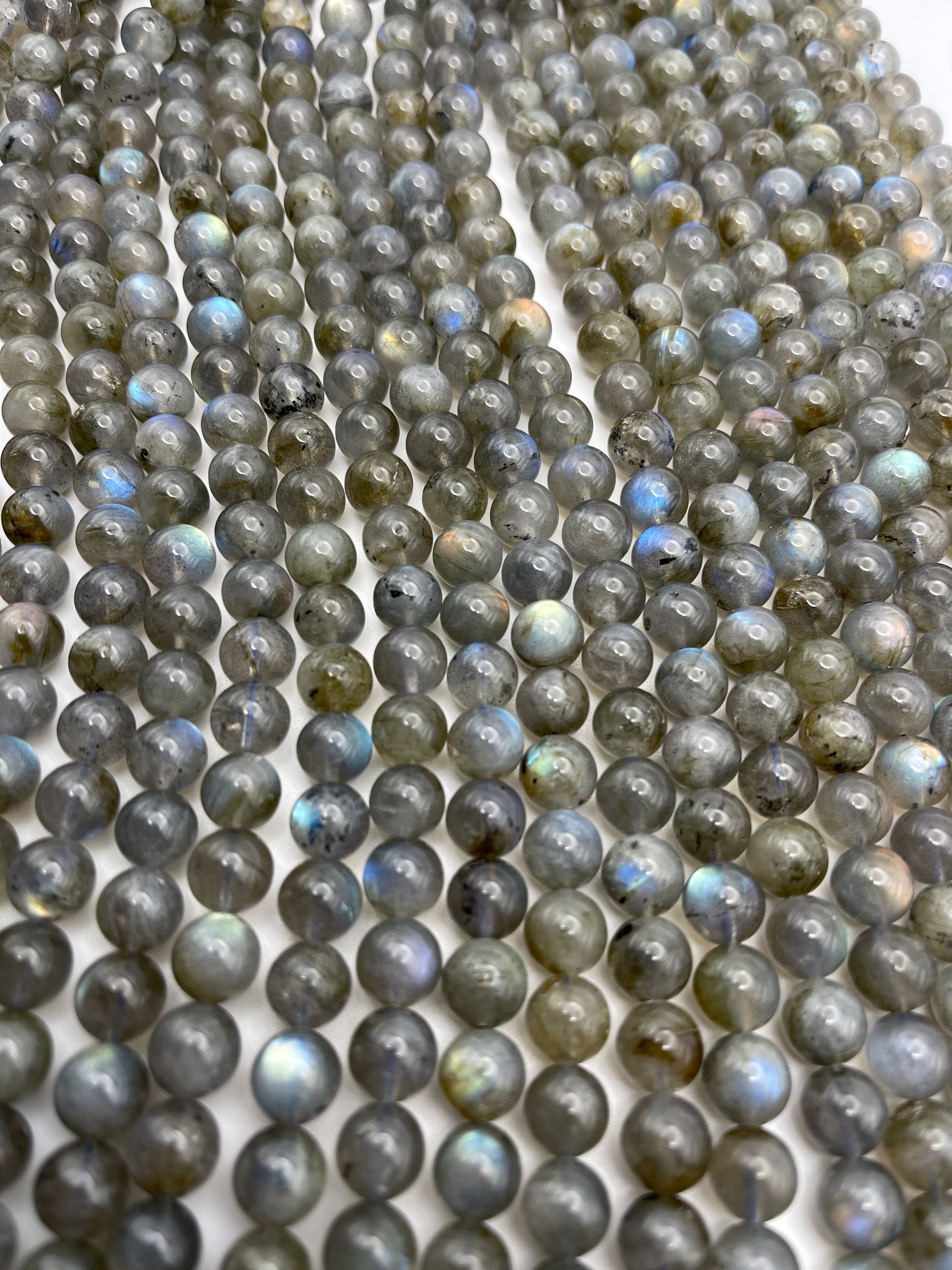Rows of 8mm Labradorite Rnd Smooth PG beads showcase a spectrum of colors from gray to blue and green on a flat surface, featuring a smooth, polished finish and a gentle play of iridescent hues.