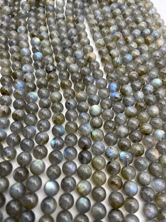 Rows of 8mm Labradorite Rnd Smooth PG beads showcase a spectrum of colors from gray to blue and green on a flat surface, featuring a smooth, polished finish and a gentle play of iridescent hues.