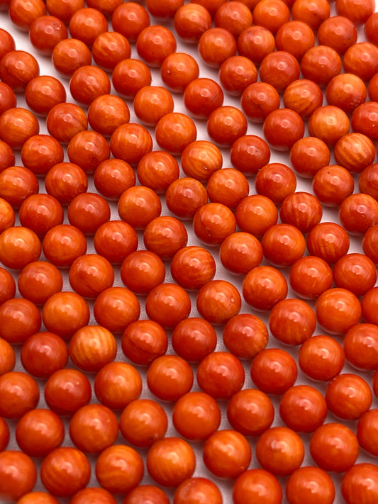 A close-up image shows numerous small, polished 6mm Round Coral beads arranged in a grid pattern. The beads have a vibrant, glossy finish and appear uniform in size, with slight variations in their color and surface texture.