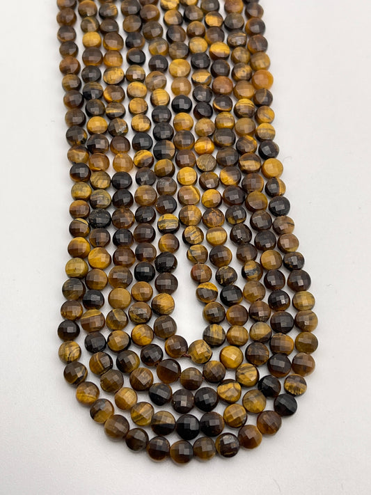 A close-up photograph of several strands of 5mm Faceted Coin Tiger's Eye beads. The beads display the stone’s characteristic golden to dark brown colors with a chatoyant effect, arranged in neat rows and lying flat against a white background.