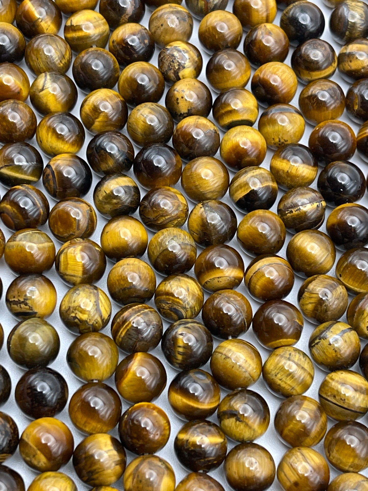 A close-up image of numerous 8mm Round Tiger's Eye gemstones neatly arranged in rows. The stones display a rich variety of golden, brown, and black bands, creating a shimmering, reflective effect across their surfaces.