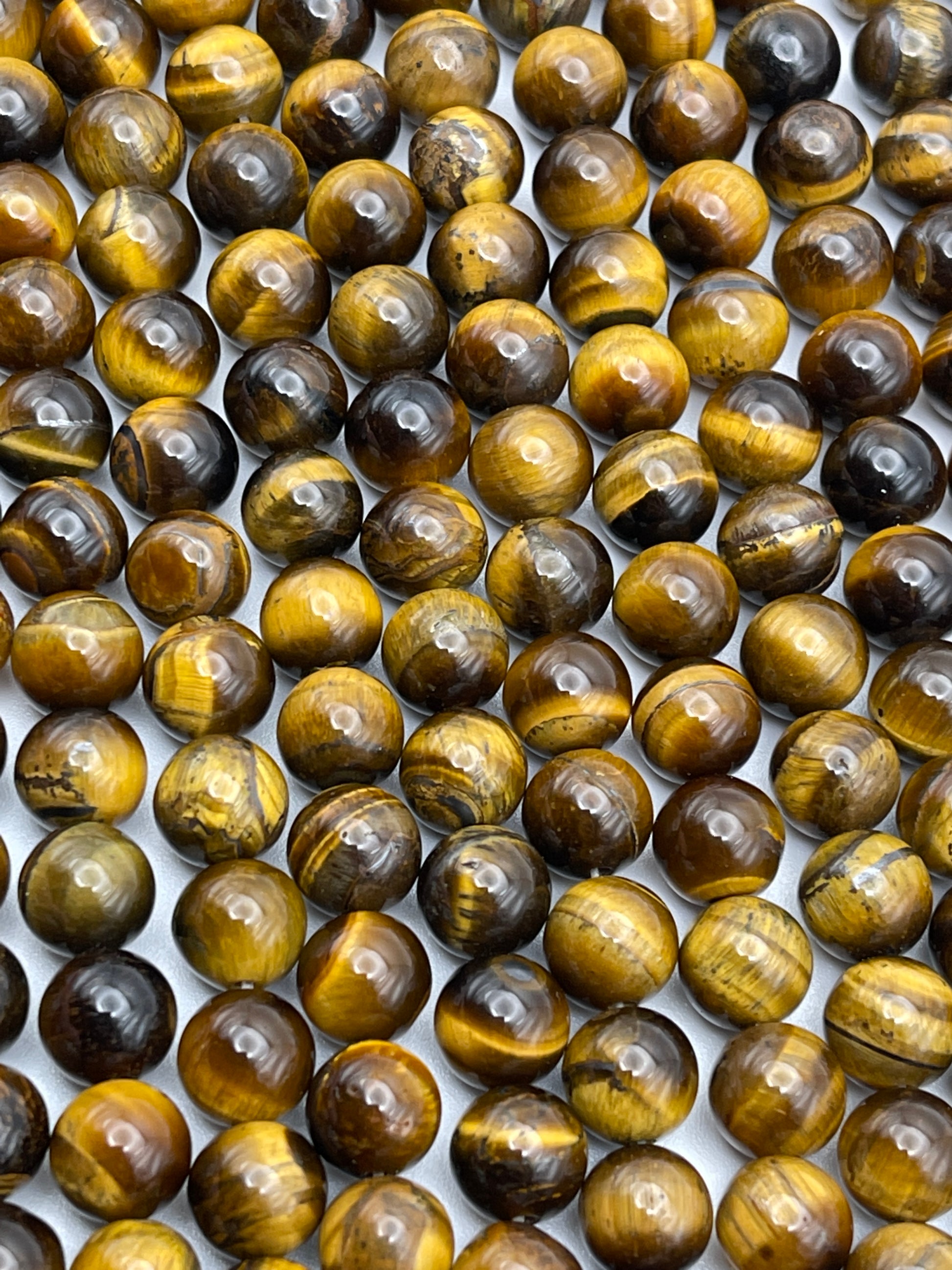 A close-up image of numerous 8mm Round Tiger's Eye gemstones neatly arranged in rows. The stones display a rich variety of golden, brown, and black bands, creating a shimmering, reflective effect across their surfaces.