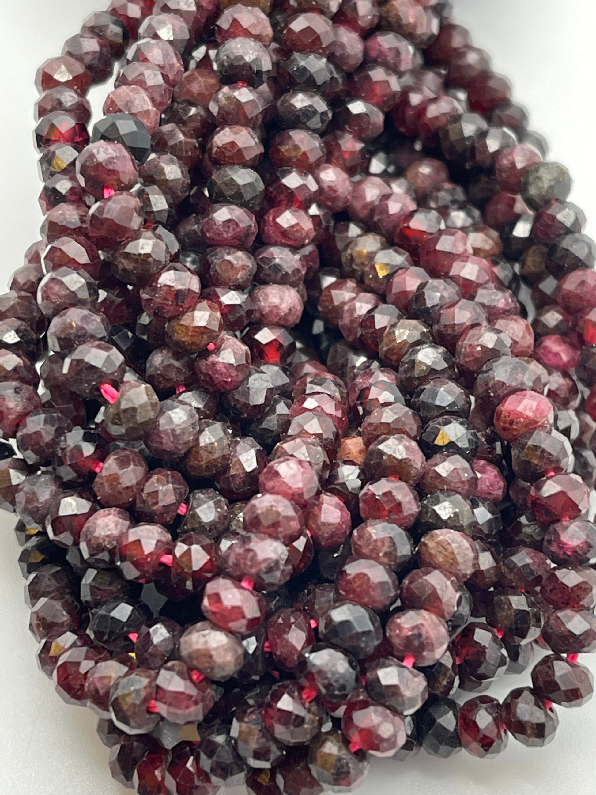 A close-up shot of a cluster of 5mm Fac Garnet Rondell beads. The beads have a glossy finish and reflect light, displaying variations in color from dark burgundy to lighter red hues. They are tightly packed and strung together, creating a rich, textured appearance.