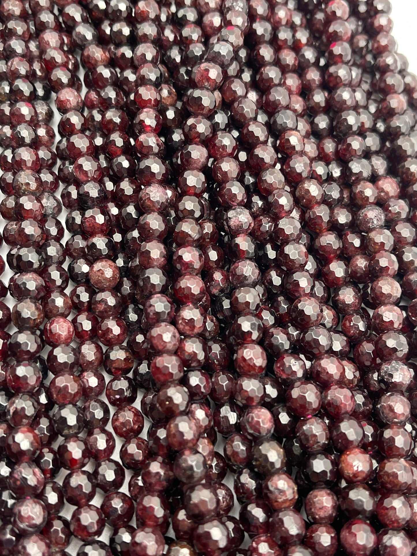 A close-up image of multiple strands of 7mm faceted garnet beads. The beads exhibit a rich, deep red hue and a polished, sparkling surface. The strands are intricately arranged, filling the entire frame.