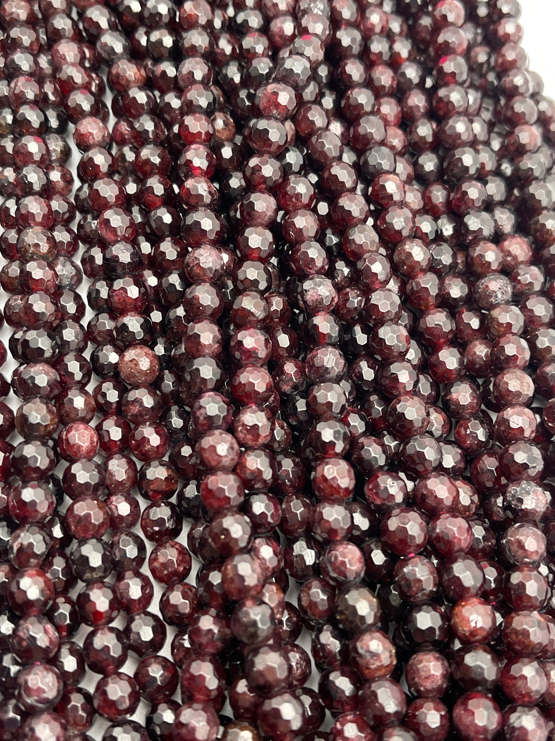 A close-up image of multiple strands of 7mm faceted garnet beads. The beads exhibit a rich, deep red hue and a polished, sparkling surface. The strands are intricately arranged, filling the entire frame.
