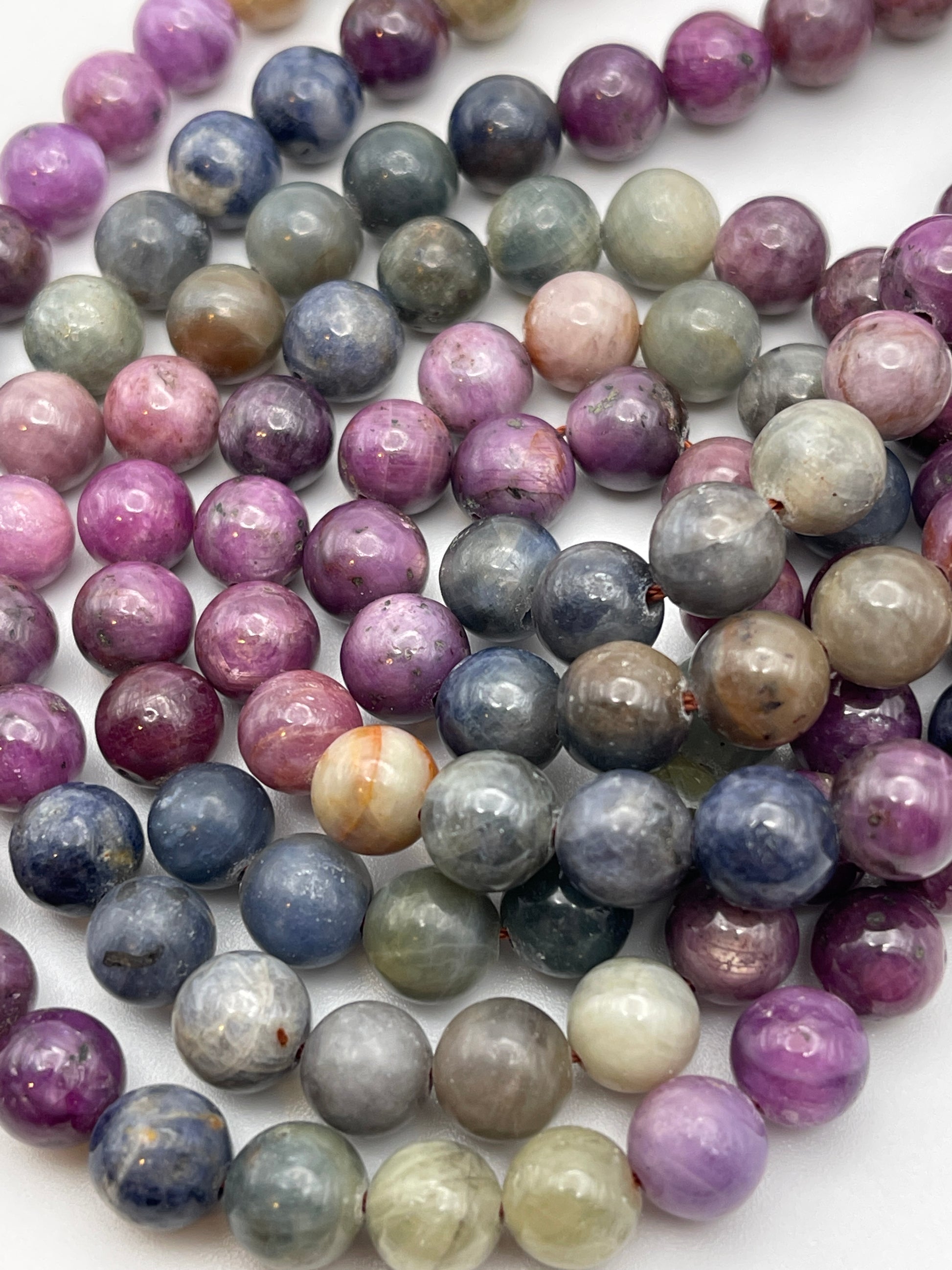 A close-up view of numerous colorful beads made from various stones. The beads, part of the 8mm Sapphire Rainbow Rnd Smooth collection, include shades of purple, green, blue, pink, and gray. They are polished and rounded, arranged closely together to showcase their smooth surfaces and natural variations.