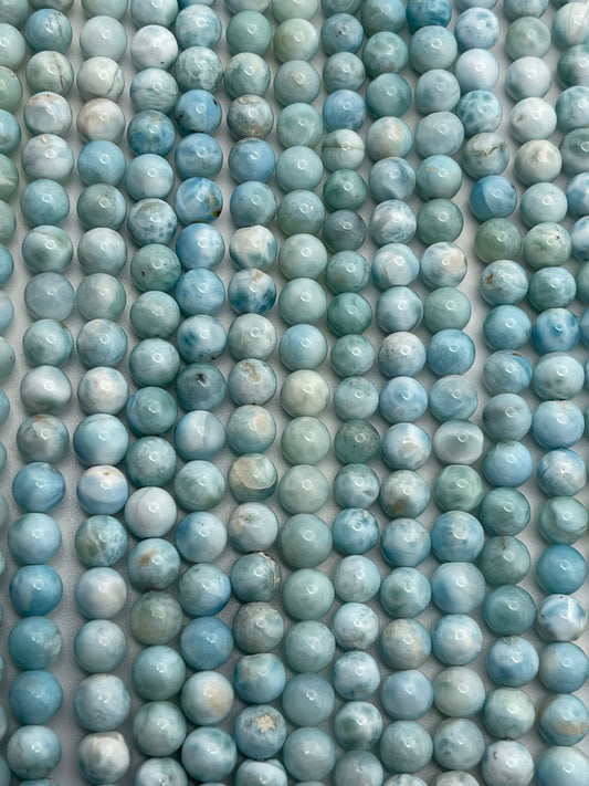 Rows of 6mm Round Larimer beads, featuring blue and turquoise hues with marbled white and grey patterns, are uniformly arranged. These beads display a spectrum of blue tones from light to deep shades, creating a calming and visually appealing texture.